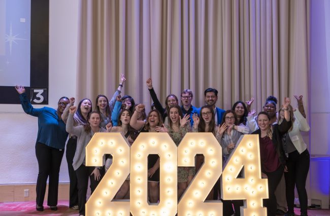 All MPH 2024 graduates standing behind2024 neon sign smiling with arms raised.