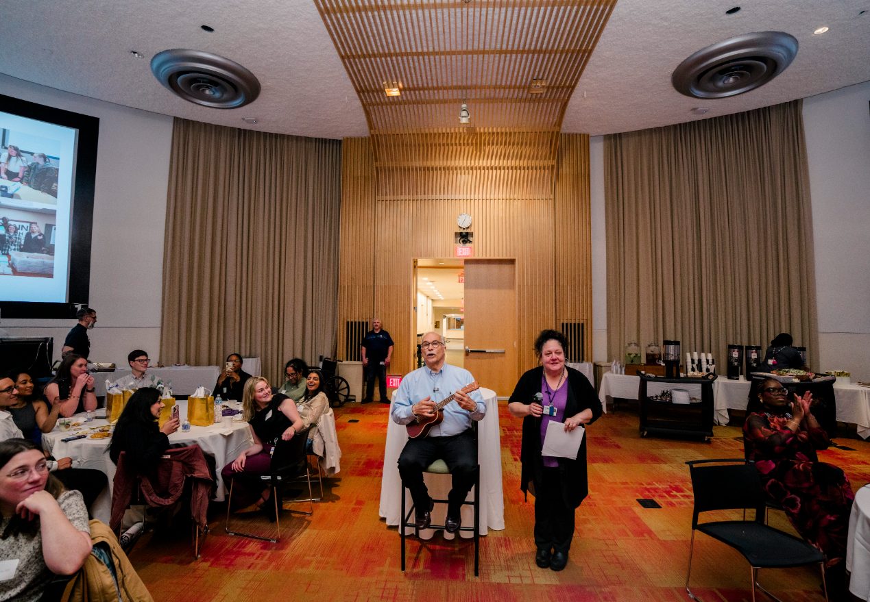 David Gregorio sits in chair holding ukulele and Lisa Parenteau stands beside him holding a microphone.